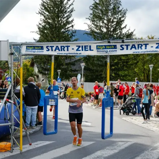 Ein Marathonläufer beendet den Wettkampf unter einem Banner, das 'Start-Ziel' steht. Zuschauer beobachten, einige stehen am Rand, andere sitzen oder stehen in der Nähe. Bäume und Berge sind im Hintergrund zu sehen.