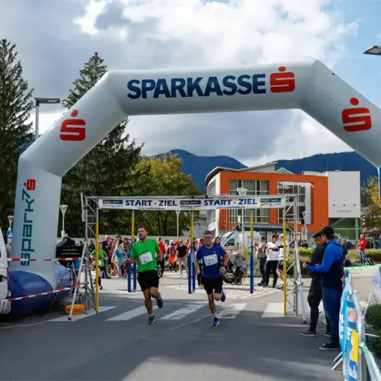 Läufer überqueren die Ziellinie eines Marathons im Freien mit einem Banner, das 'Sparkasse Start Ziel' lautet.