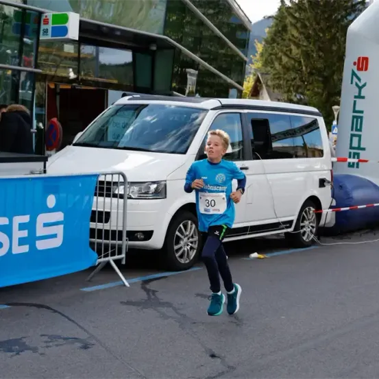 Ein junger Junge läuft einen Marathon und trägt ein blaues Shirt und Turnschuhe. Er läuft an einem weißen Van mit der Nummer 30 vorbei. Ein blauer Banner befindet sich in der Nähe.