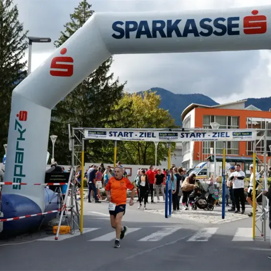 Ein Marathonläufer überquert die Ziellinie unter einem weißen Bogen mit dem Wort Sparkasse. Zuschauer und Freiwillige stehen hinter ihm, einige halten Plakate. Im Hintergrund befinden sich Gebäude und eine Bergkette.