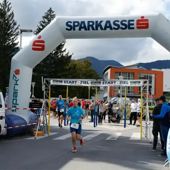 Läufer 46 überquert die Ziellinie eines Marathonrennens. Zuschauer sind versammelt. Bäume und Berge sind im Hintergrund.