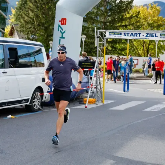 Ein Mann läuft bei einem Marathon, hinter ihm parkt ein weißer Van. Es gibt ein Banner mit der Aufschrift Start-Ziel. Zuschauer beobachten von der Seite.