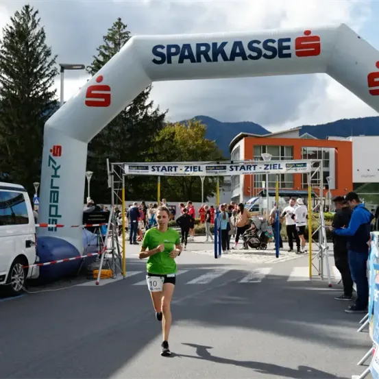 Eine Läuferin in einem grünen Shirt und schwarzen Shorts läuft unter einem weißen Bogen mit dem Wort 'Sparkasse' darauf. Menschen stehen und beobachten. Links steht ein Van und in der Ferne ein Kinderwagen.