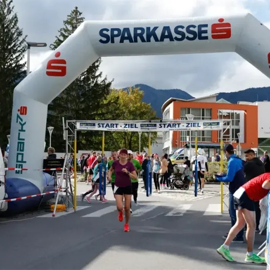 Ein vom Sparkasse gesponserter Marathon mit Läufern, die unter einem Banner hindurchlaufen. Zuschauer, Bäume und Gebäude im Hintergrund.