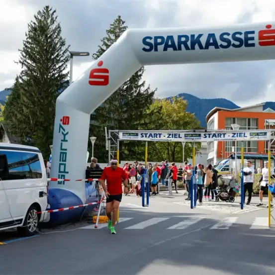 Ein Marathonläufer läuft unter einem 'Sparkasse'-Banner. Zuschauer beobachten hinter Absperrungen. Die Szene umfasst Bäume, Berge und einen geparkten weißen Van in der Nähe.