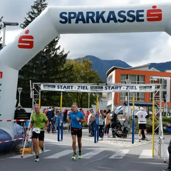 Zwei Männer laufen bei einem Marathon mit einem 'Start-Ziel'-Bogen und einem großen 'SPARKASSE'-Banner. Zuschauer und Freiwillige sind anwesend, mit einem orangefarbenen Gebäude im Hintergrund.