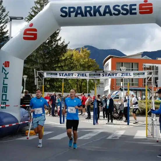 Zwei Läufer nehmen an einem Marathon teil, mit einem weißen Bogen, der 'SPARKASSE' am Start anzeigt. Zuschauer und Freiwillige säumen die Strecke, mit einem Gebäude im Hintergrund und Bäumen.