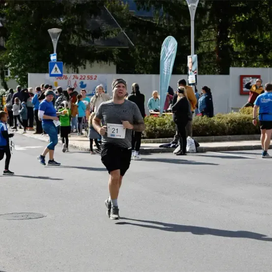 Ein Mann in einem grauen Shirt mit der Nummer 21 läuft einen Marathon. Zuschauer stehen am Straßenrand, einige fotografieren. In der Ferne sind Straßenlaternen und ein Schild.
