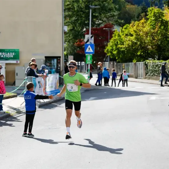 Ein Marathonläufer in einem grünen Shirt und schwarzen Shorts erhält eine Wasserflasche von einem jungen Jungen in einem blauen Shirt. Der Junge steht auf dem Bürgersteig, und Zuschauer beobachten von einer grünen Absperrung aus.