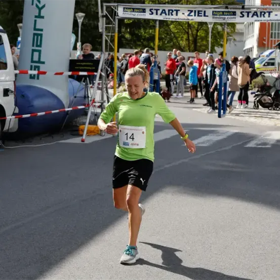 Ein Marathonläufer mit der Nummer 14 auf dem Trikot läuft auf der Straße. Dahinter steht eine Menschenmenge und ein Kinderwagen. Der Lauf hat ein Banner mit dem Text 'Start-Ziel'.