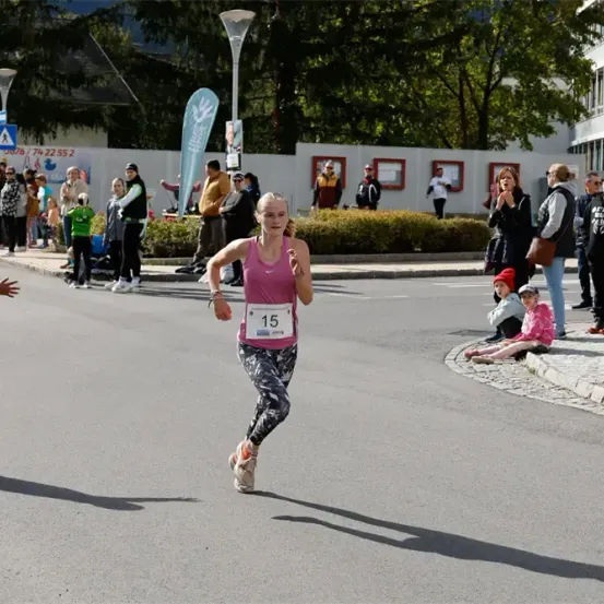 Eine Frau mit einem Nummer 15 gekennzeichneten Startnummern-Bib läuft auf der Straße, während Leute vom Bürgersteig zusehen.
