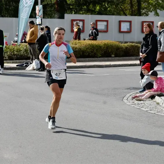 Eine Frau läuft einen Marathon und trägt ein Trikot mit der Nummer 47. Zuschauer stehen und sitzen am Straßenrand und beobachten sie.
