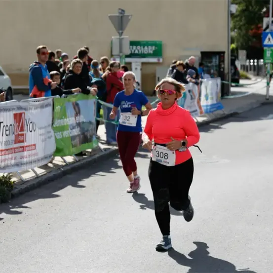 Zwei Frauen laufen einen Marathon, eine trägt ein rotes Shirt, die andere blau. Zuschauer stehen hinter einem Banner am Straßenrand.