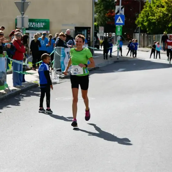 Eine Frau in Grün läuft einen Marathon. Ein junger Junge in Blau steht hinter ihr. Zuschauer jubeln vom Bürgersteig.