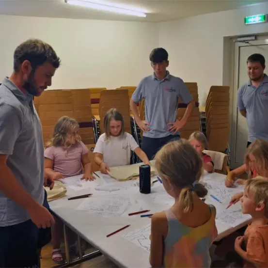 Eine Gruppe von Kindern und zwei Männern sitzen um einen Tisch herum und zeichnen mit Bleistiften. Die Männer stehen hinter dem Tisch, einer trägt ein graues Poloshirt und der andere ein blaues Poloshirt.