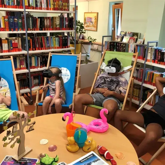 Vier Kinder sitzen in Stühlen in einer Bibliothek und tragen VR-Headsets. Ein Tisch hat Spielzeug, Bücher und einen Baum. Regale voller Bücher stehen hinter ihnen.