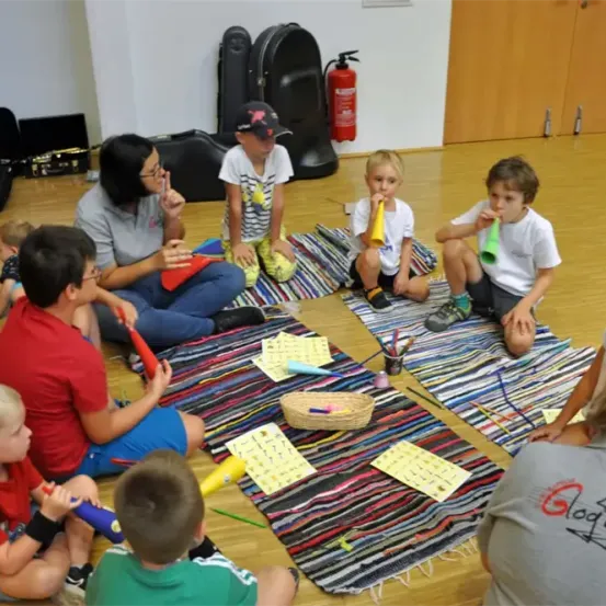 Eine Gruppe von Kindern sitzt im Kreis auf dem Boden und hält Musikinstrumente in den Händen. Eine erwachsene Frau sitzt unter ihnen. Der Boden ist aus Holz und mehrere Musikinstrumente und Papiere sind umher verstreut. Im Hintergrund befinden sich ein Feuerlöscher und einige schwarze Koffer.