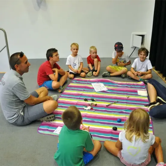 Eine Gruppe von Kindern und Erwachsenen sitzt um eine bunte Decke herum und beschäftigt sich mit einer Aktivität mit Spielzeug und einem weißen Papier.
