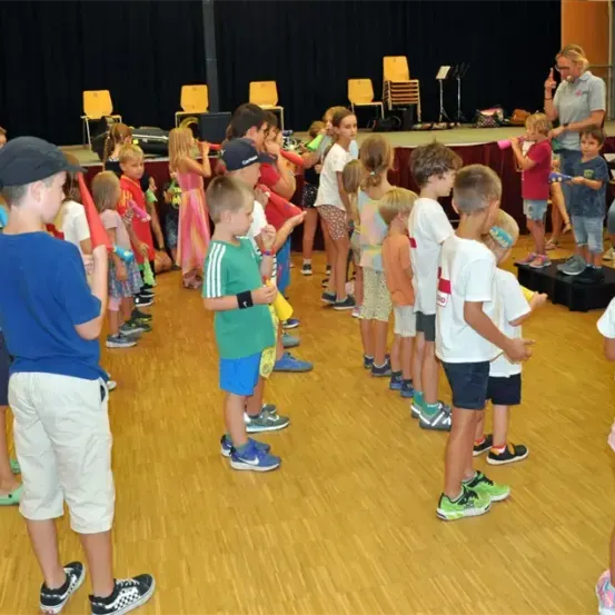 Eine Gruppe von Kindern steht in einer Reihe auf einer Bühne, trägt Turnschuhe und einige Hüte. Ein Erwachsener steht vorne, vielleicht um sie zu unterrichten. Hinter ihnen sind Stühle und eine Bühne mit schwarzem Hintergrund.