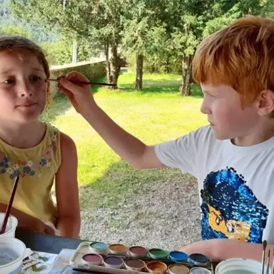 Ein Junge und ein Mädchen malen im Freien an einem sonnigen Tag Gesichter. Das Mädchen hat sein Gesicht bemalt, während der Junge einen Pinsel hält. Sie sitzen an einem Tisch mit Malutensilien.
