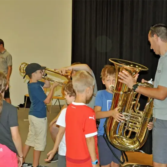 Eine Gruppe von Kindern und einem Erwachsenen spielt Blechblasinstrumente, darunter Trompeten und Tuben, in einem Raum mit einem schwarzen Vorhang im Hintergrund.