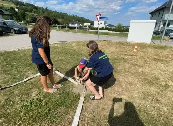 Drei Personen arbeiten auf einer Grasfläche, wahrscheinlich einer Baustelle. Zwei Frauen und ein Kind. Die Frau auf der linken Seite steht, während die andere Frau kniet. Ein Schlauch liegt auf dem Boden.