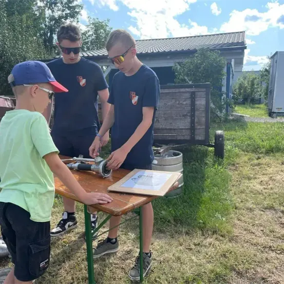 Drei Jungen arbeiten an einem Gerät auf einem Tisch im Freien und tragen eine Sonnenbrille. Dahinter sind ein Haus und Fässer zu sehen.