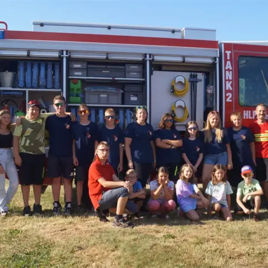 Eine Gruppe von Menschen, darunter Erwachsene und Kinder, posiert für ein Foto vor einem roten Feuerwehrauto. Das Auto ist offen und zeigt verschiedene Ausrüstungsgegenstände im Inneren.