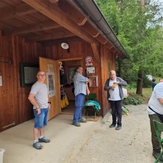 Drei Männer stehen vor einer Holzhütte mit offener Tür, ein weiterer Mann sitzt auf einem Stuhl in der Nähe. Bäume und Grünfläche umgeben das Gebiet.