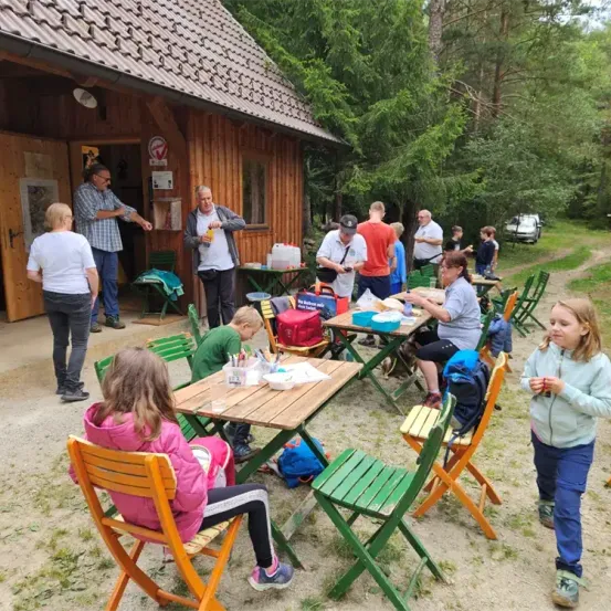 Eine Gruppe von Menschen versammelt sich um Tische vor einem Holzgebäude mit Bäumen im Hintergrund. Einige sitzen, andere stehen, und ein Auto ist in der Nähe geparkt.