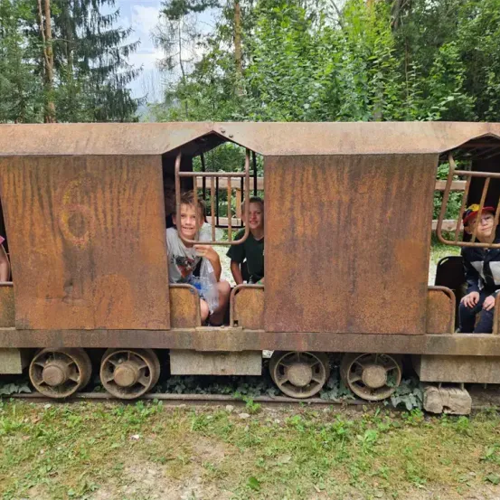 Vier Kinder sitzen in einem rostigen Zugwaggon und schauen aus den Fenstern. Der Zug steht auf einem grasbewachsenen Gleis, umgeben von Bäumen.