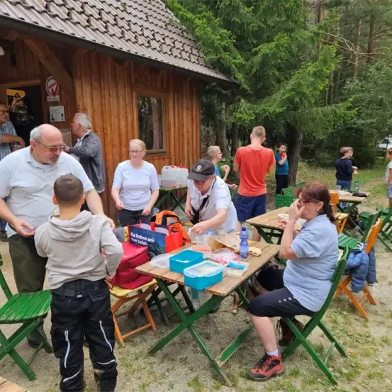 Eine Gruppe von Menschen versammelt sich um Tische in einem bewaldeten Gebiet. Einige essen, andere stehen und unterhalten sich. Ein Gebäude mit einem Schild ist im Hintergrund.