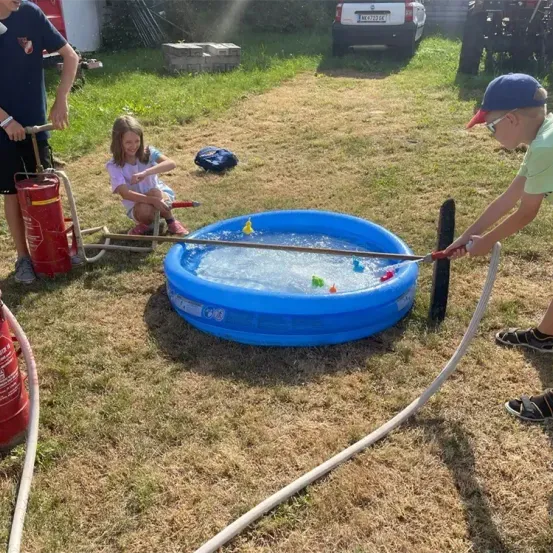 Eine Gruppe von Kindern spielt mit einem Wasserschlauch und füllt einen kleinen blauen Pool mit Wasser. Ein Junge hält einen Schlauch, während ein Mädchen in der Nähe sitzt.