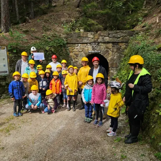 Eine Gruppe von Kindern und Erwachsenen in gelben Schutzhelmen und Regenmänteln steht vor einem steinernen Tunneleingang in einem Wald.