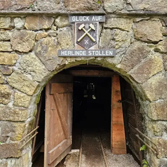 Der Eingang zu Herlinde Stollen, einem Bergwerk, zeigt eine Steinmauer mit einem Schild, auf dem 'Gluck Auf' und 'Herlinde Stollen' steht. Eine Holztür ist offen und zeigt den dunklen Innenraum.