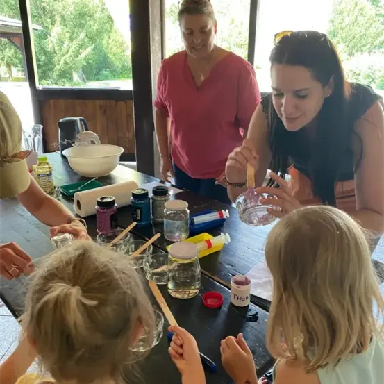 Eine Gruppe von Frauen und Kindern arbeitet gemeinsam an einem Wissenschaftsprojekt. Sie mischen Flüssigkeiten in Bechern und Gläsern, wobei eine Frau sie anleitet. Der Tisch ist mit verschiedenen Flaschen und Werkzeugen übersät.