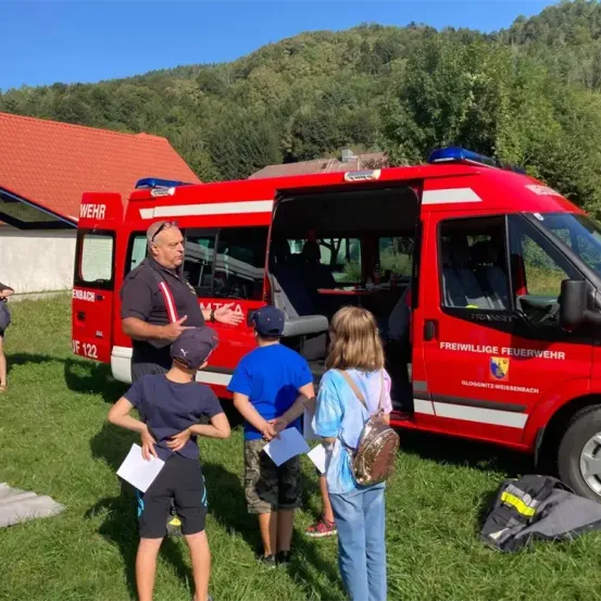Ein Mann spricht mit drei Kindern vor einem roten Feuerwehrwagen an einem sonnigen Tag.