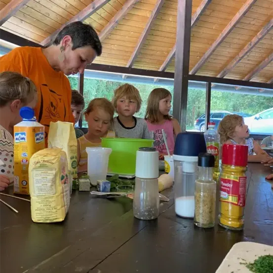 Eine Gruppe von Kindern und einem Erwachsenen versammelt sich um einen Tisch mit verschiedenen Zutaten. Flaschen, ein gelber Beutel und eine grüne Schüssel stehen auf dem Tisch. Der Erwachsene scheint die Kinder zu unterweisen.