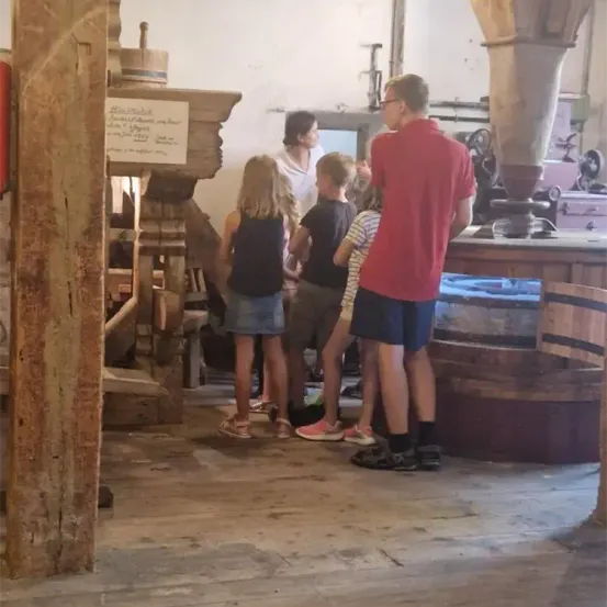 Eine Gruppe von Kindern und Erwachsenen steht in einem rustikalen Raum, möglicherweise einem Museum, und beobachtet eine alte Holzstruktur mit einem Schild. In der Nähe ist eine große Maschine zu sehen.