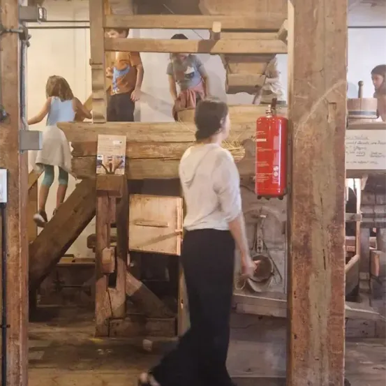 Eine Gruppe von Menschen befindet sich in einer Holzstruktur, wahrscheinlich einer alten Mühle. Einige sind auf den Treppen, andere in der Nähe des Mühlsteins. Ein Feuerlöscher ist an der Wand montiert.