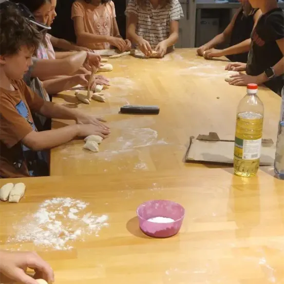 Kinder an einem Tisch rollen Teig zu Formen. Ein kleiner Teller mit Mehl und eine Ölflasche sind in der Nähe.