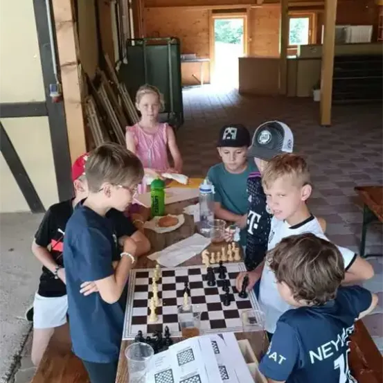 Mehrere Kinder spielen Schach auf einem Tisch in einem Holzgebäude. Ein Mädchen steht in der Nähe und beobachtet.