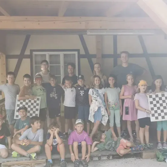 Eine Gruppe von Kindern und Erwachsenen posiert für ein Foto in einem Raum mit Holzbalken. Einige halten Schachbretter, und es stehen und sitzen mehrere auf dem Boden.