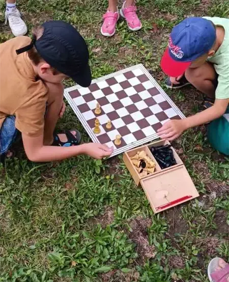 Zwei Jungen spielen Schach auf einem Grasfeld. Ein Junge trägt ein braunes Shirt, der andere ein grünes.