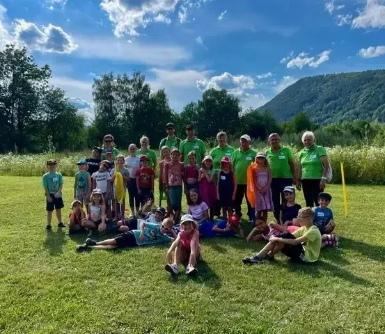 Eine Gruppe von Kindern und Erwachsenen in grünen Shirts posiert für ein Foto auf einer Wiese, mit Bergen im Hintergrund.