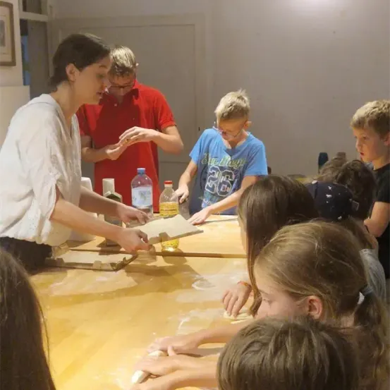 Eine Frau in einem weißen Hemd zeigt Kindern, wie man Teig auf einem Holztisch ausrollt. Zwei Erwachsene, einer in Rot und einer in Blau, stehen hinter den Kindern.