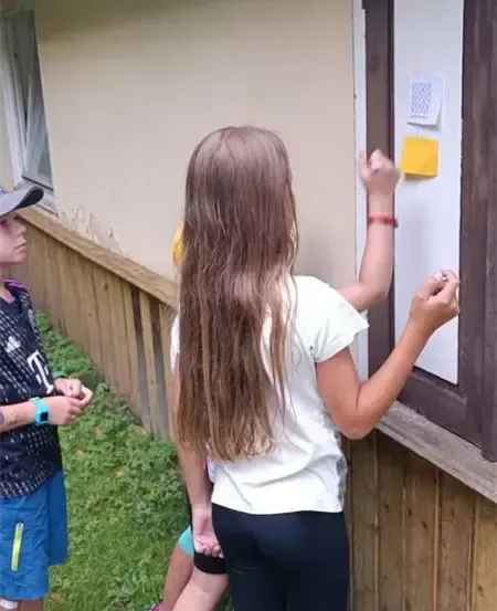 Drei Kinder stehen vor einem Gebäude, zwei Mädchen vor einer Tür mit Notizen und ein Junge, der sie ansieht. Die Mädchen haben lange Haare und tragen Armbänder, und der Junge trägt eine Mütze.