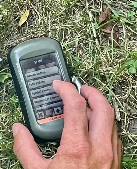 Eine Hand hält ein graues Garmin GPS-Gerät und zeigt ein Menü auf dem Bildschirm mit Optionen wie 'Namen ändern' und 'Foto ändern'. Das Gerät liegt auf Gras.