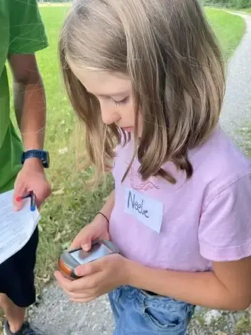 Ein Mädchen mit blonden Haaren benutzt ein Handheld-GPS-Gerät und schaut auf ein Stück Papier hinunter.