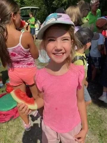 Ein junges Mädchen in einem rosa Shirt und Hut hält ein Stück Wassermelone. Hinter ihr stehen und sitzen mehrere Kinder und Erwachsene auf dem Gras. Einige tragen Hüte und Turnschuhe.